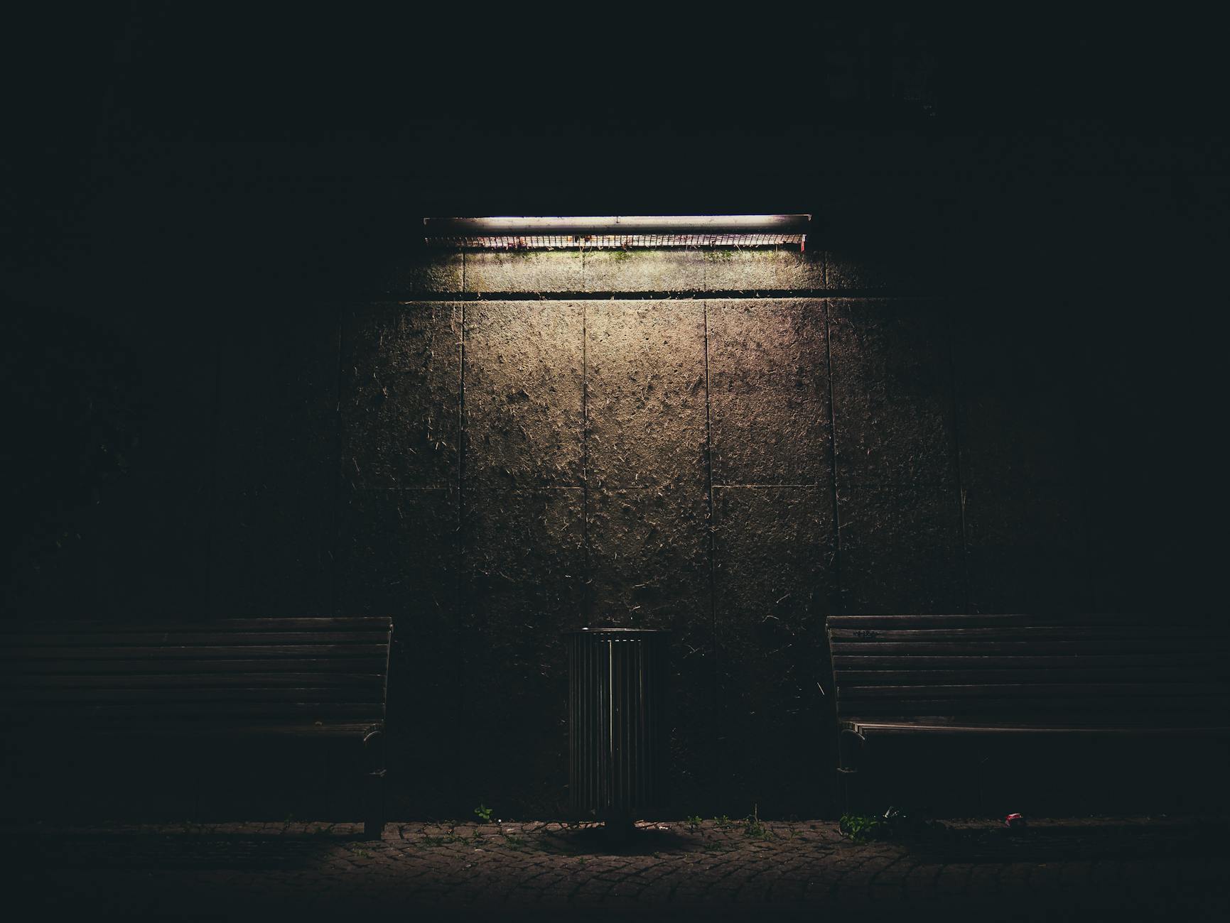 Dark urban night scene with benches and a fluorescent lamp casting soft light on the wall.