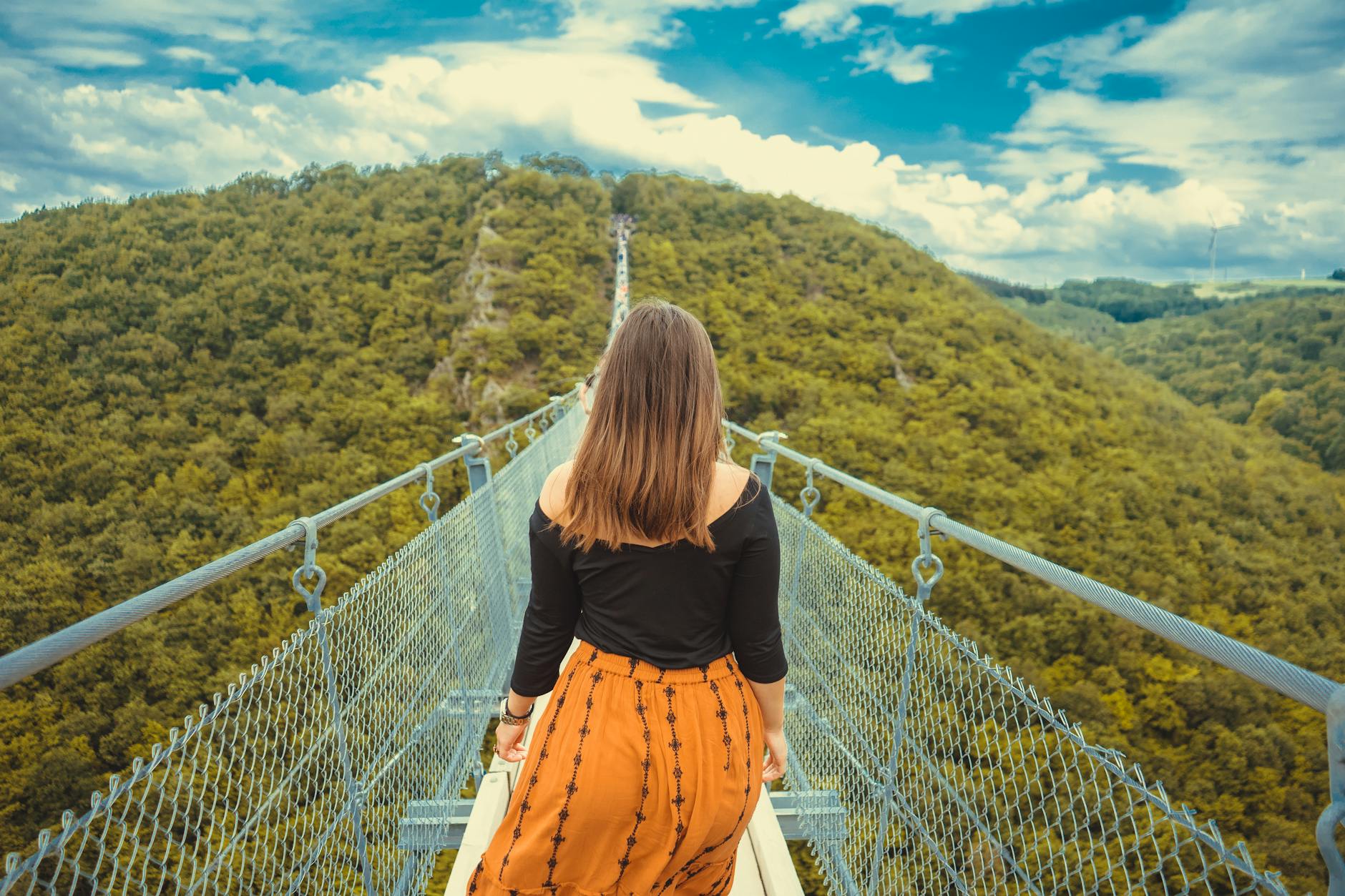 A woman walks along a canopy bridge set amidst a lush green forest and scenic hills.