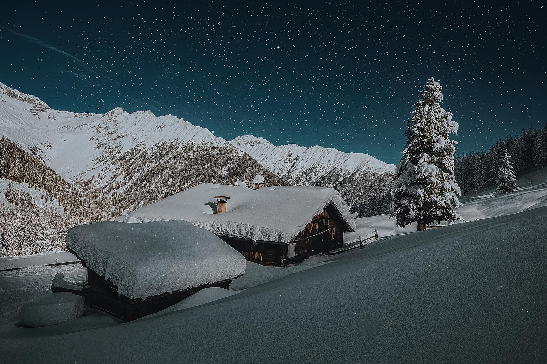 A serene winter night with cabins blanketed in snow, beneath an expansive starry sky and snow-capped mountains.