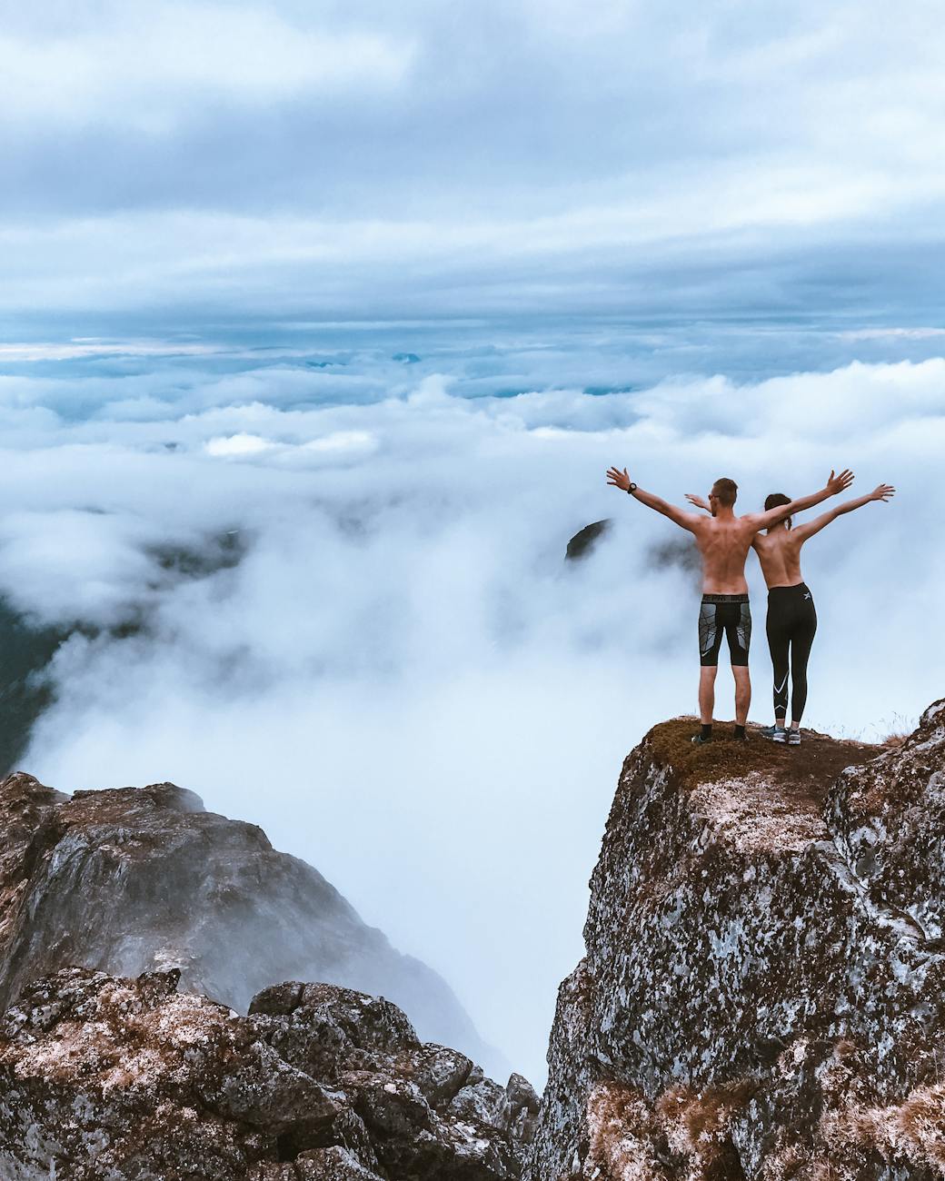 Two hikers stand on a rocky cliff, embracing the expansive mountain view and clouds, symbolizing freedom and exploration. Two hikers stand on a rocky cliff, embracing the expansive mountain view and clouds, symbolizing freedom and exploration.