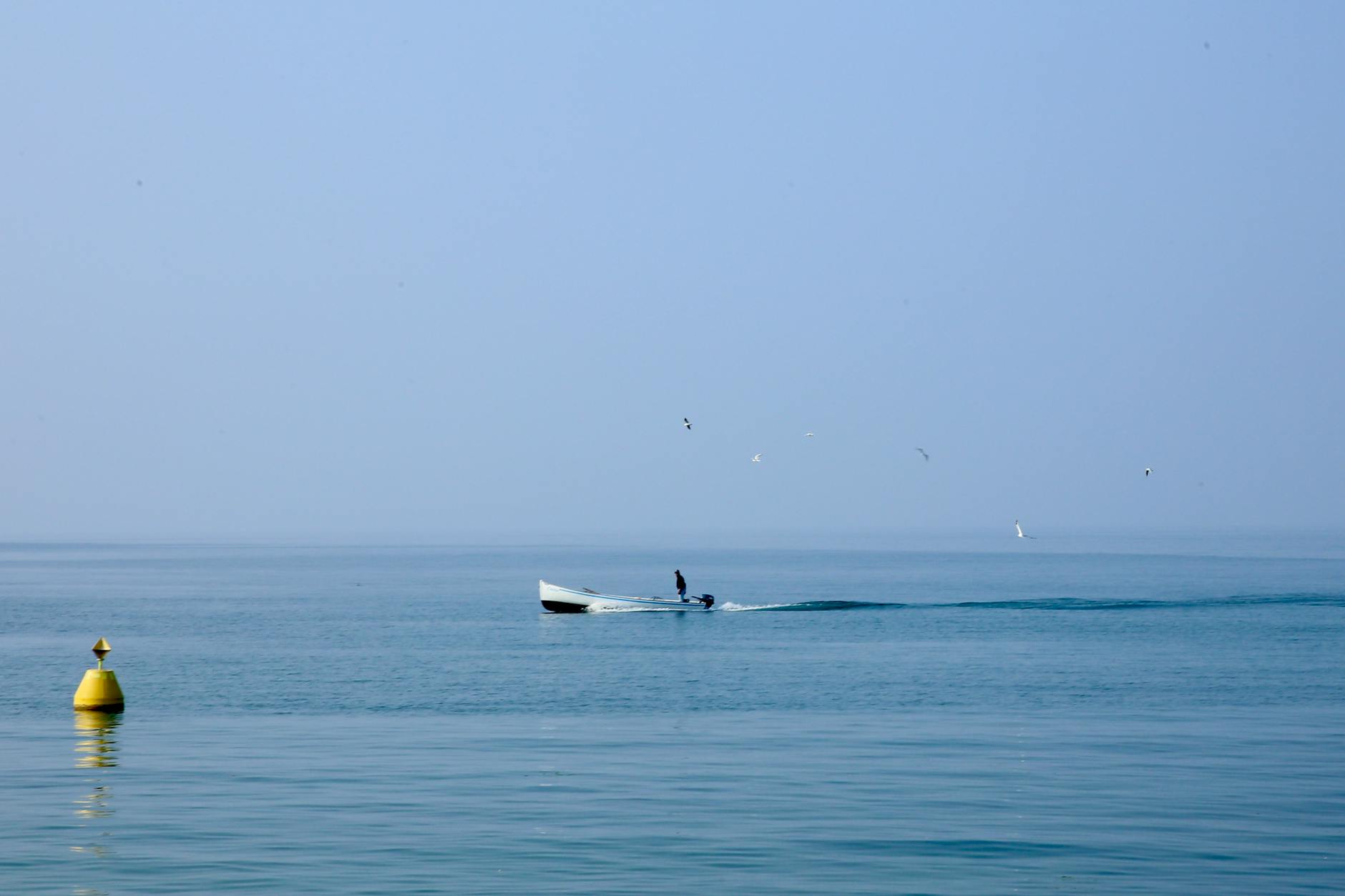 A lone figure rides a small boat across a calm, vast ocean under clear blue skies. A lone figure rides a small boat across a calm, vast ocean under clear blue skies.
