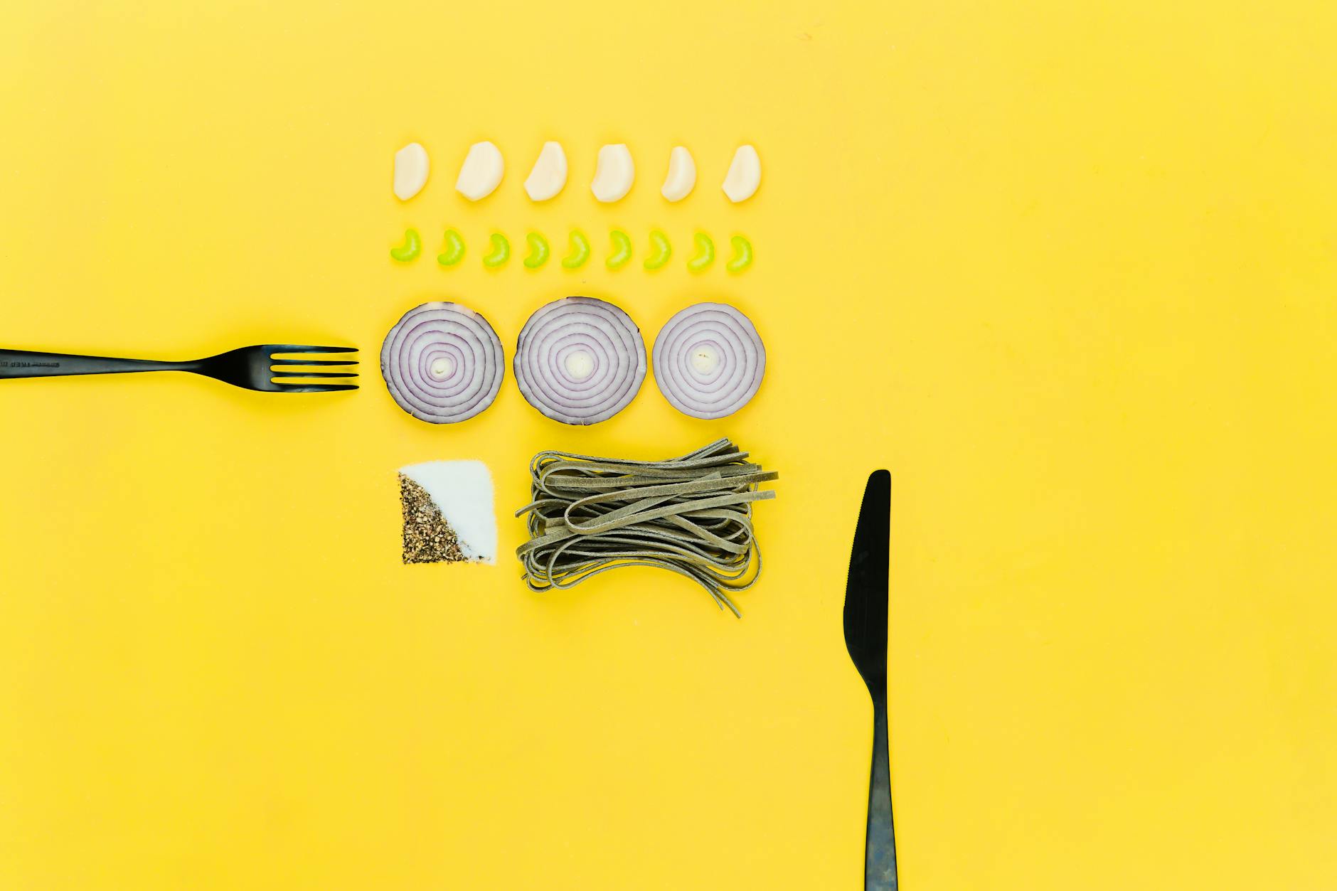 A colorful flatlay of pasta, onion, garlic, herbs, and cutlery on a yellow background. A colorful flatlay of pasta, onion, garlic, herbs, and cutlery on a yellow background.