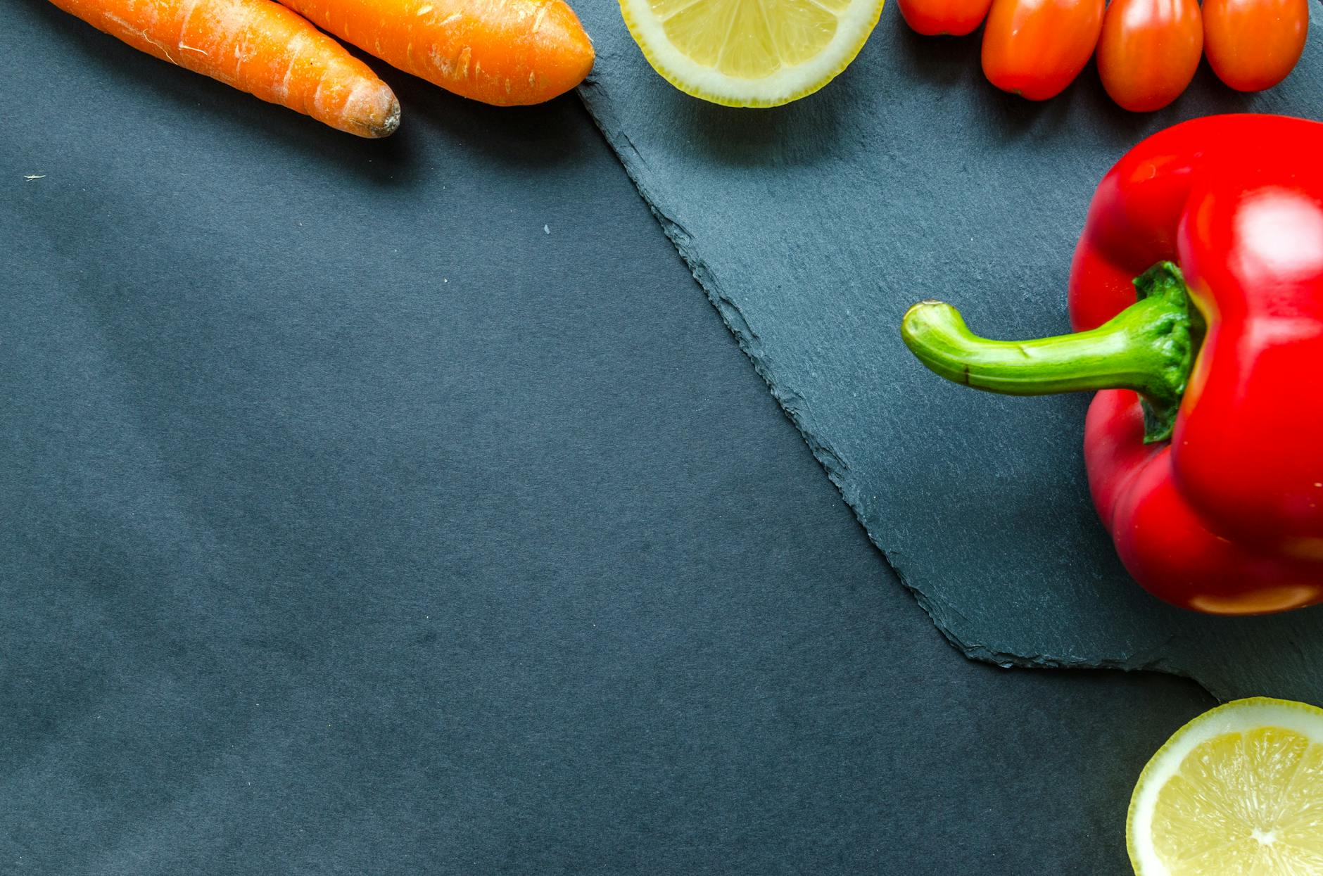 A vibrant assortment of fresh vegetables and fruits including red bell pepper and carrots on a slate surface. A vibrant assortment of fresh vegetables and fruits including red bell pepper and carrots on a slate surface.