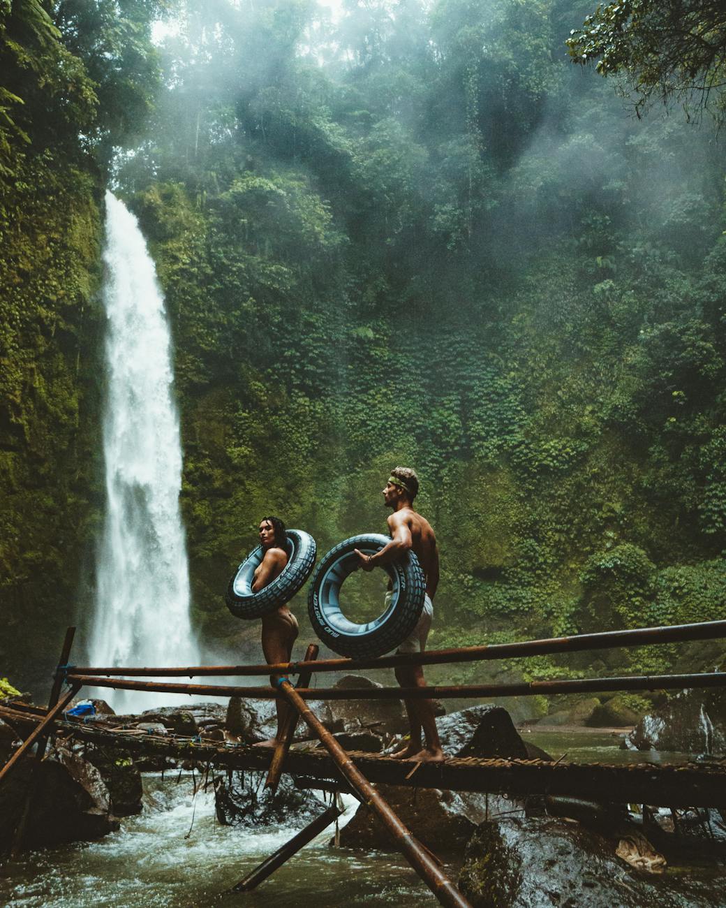 A couple in swimwear with float tubes exploring a lush waterfall in Bali, Indonesia.
