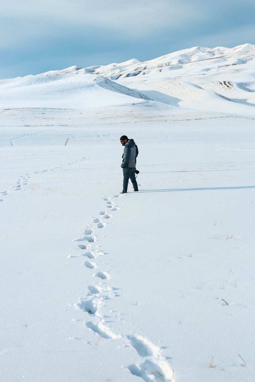 A lone man walks in a vast snowy field with distant mountains under a clear sky.