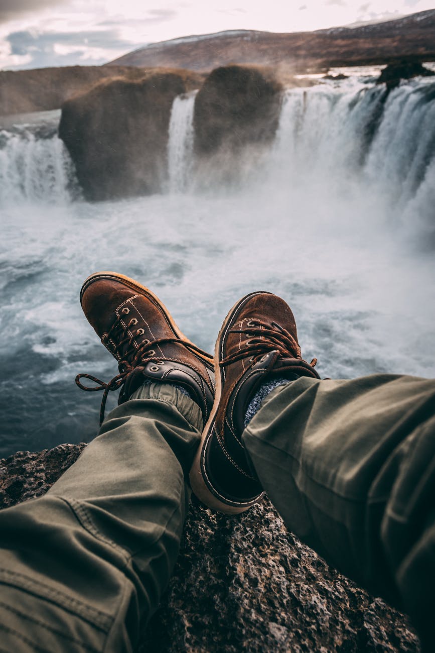 Person in hiking boots sitting at cliff’s edge, overlooking breathtaking waterfall.