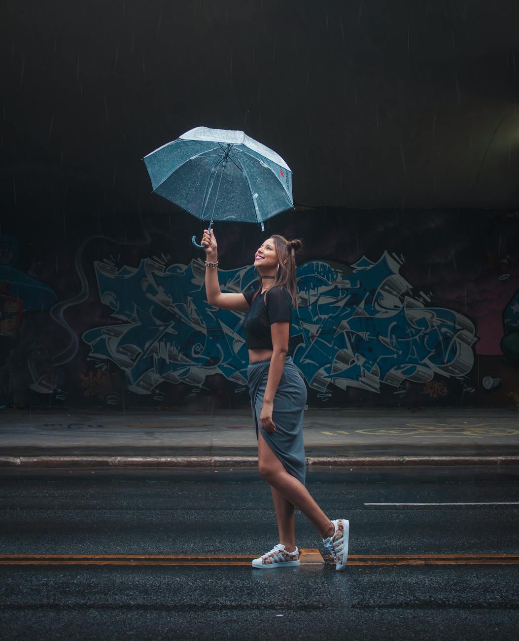 Stylish woman enjoying rain under umbrella with graffiti backdrop.