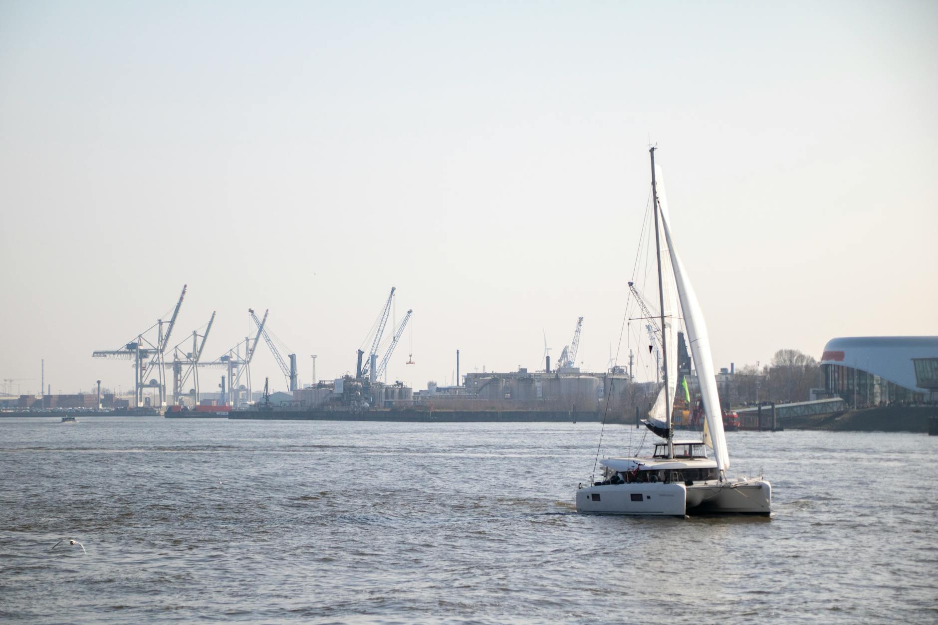 A sailboat cruises on the Elbe River with Hamburg’s port cranes in the background.