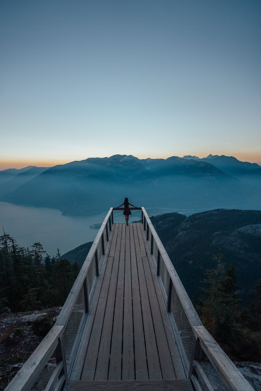 A lone person stands on a wooden boardwalk, gazing into the foggy mountain range, under a clear sky at dawn.