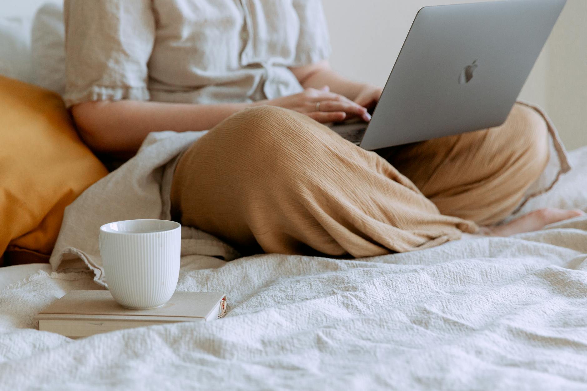 Woman enjoying downtime, using laptop on bed with a coffee cup nearby, in a relaxed home setting.