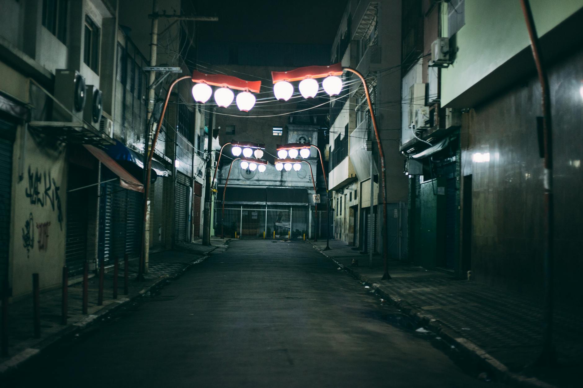 A deserted urban alley at night featuring unique red-lit hanging lights, creating a dramatic atmosphere.