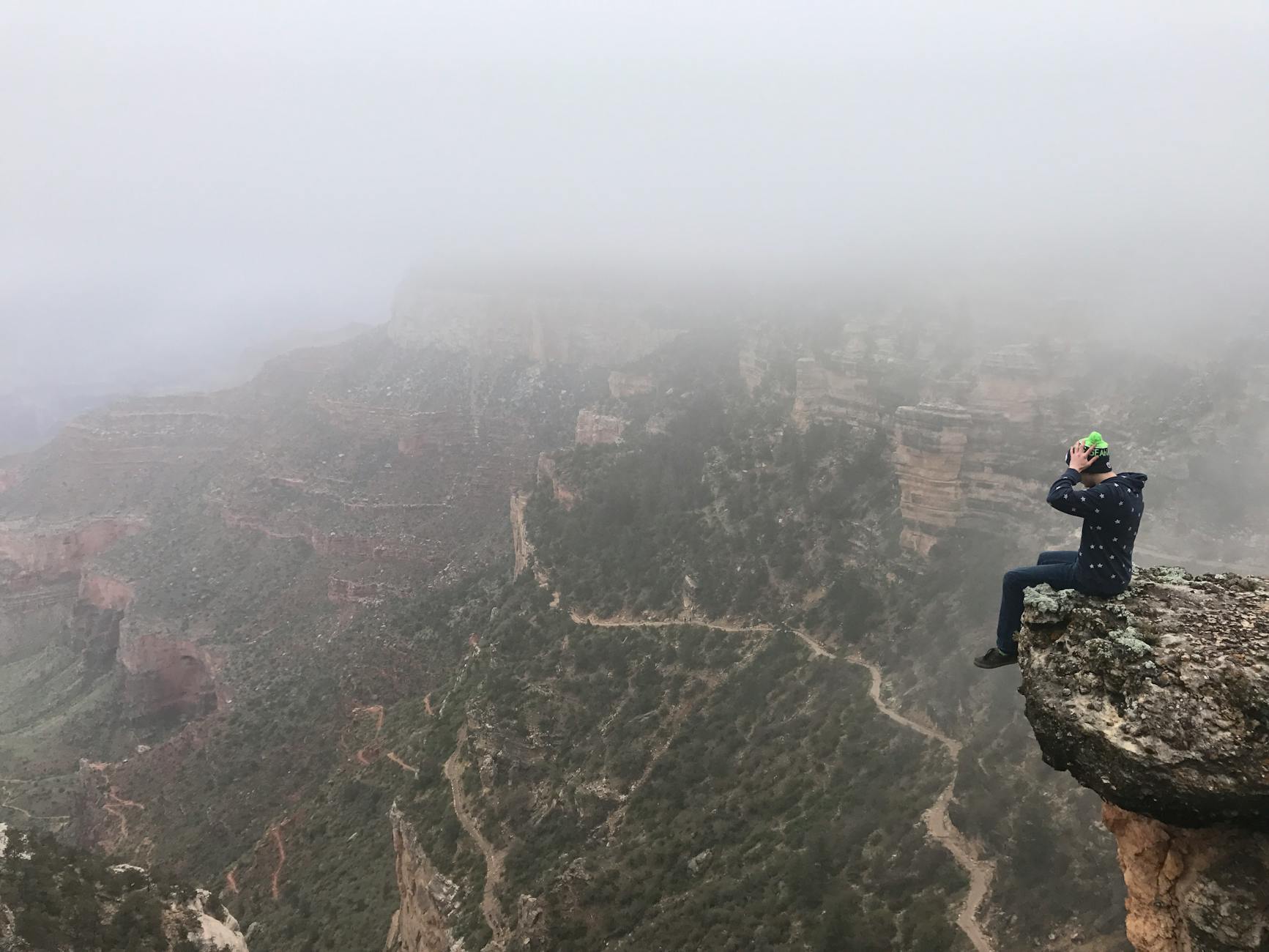 A daring hiker sits on a foggy cliff edge of the Grand Canyon, capturing the serene magnitude of nature.