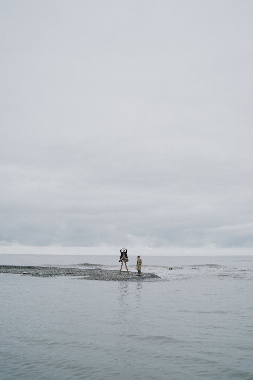 Two people stand on a small sandy beach against a gloomy, overcast seaside backdrop.