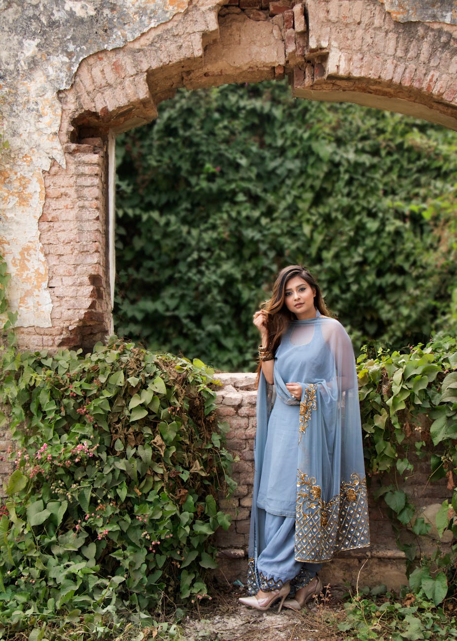 A woman in a beautiful blue traditional dress poses elegantly by ivy-covered ruins, exuding grace and charm.