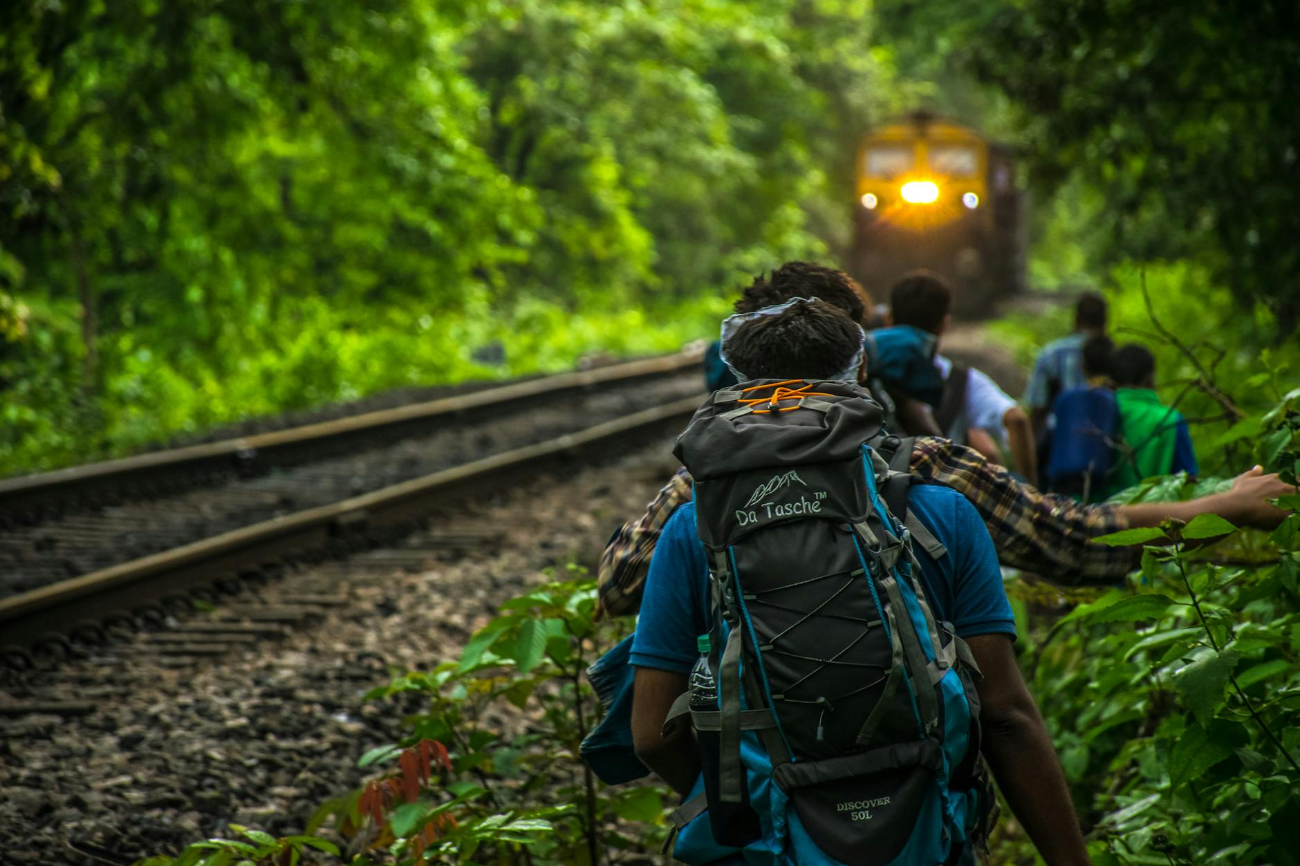 Group of backpackers walking along railway tracks through lush forest with approaching train. Group of backpackers walking along railway tracks through lush forest with approaching train.