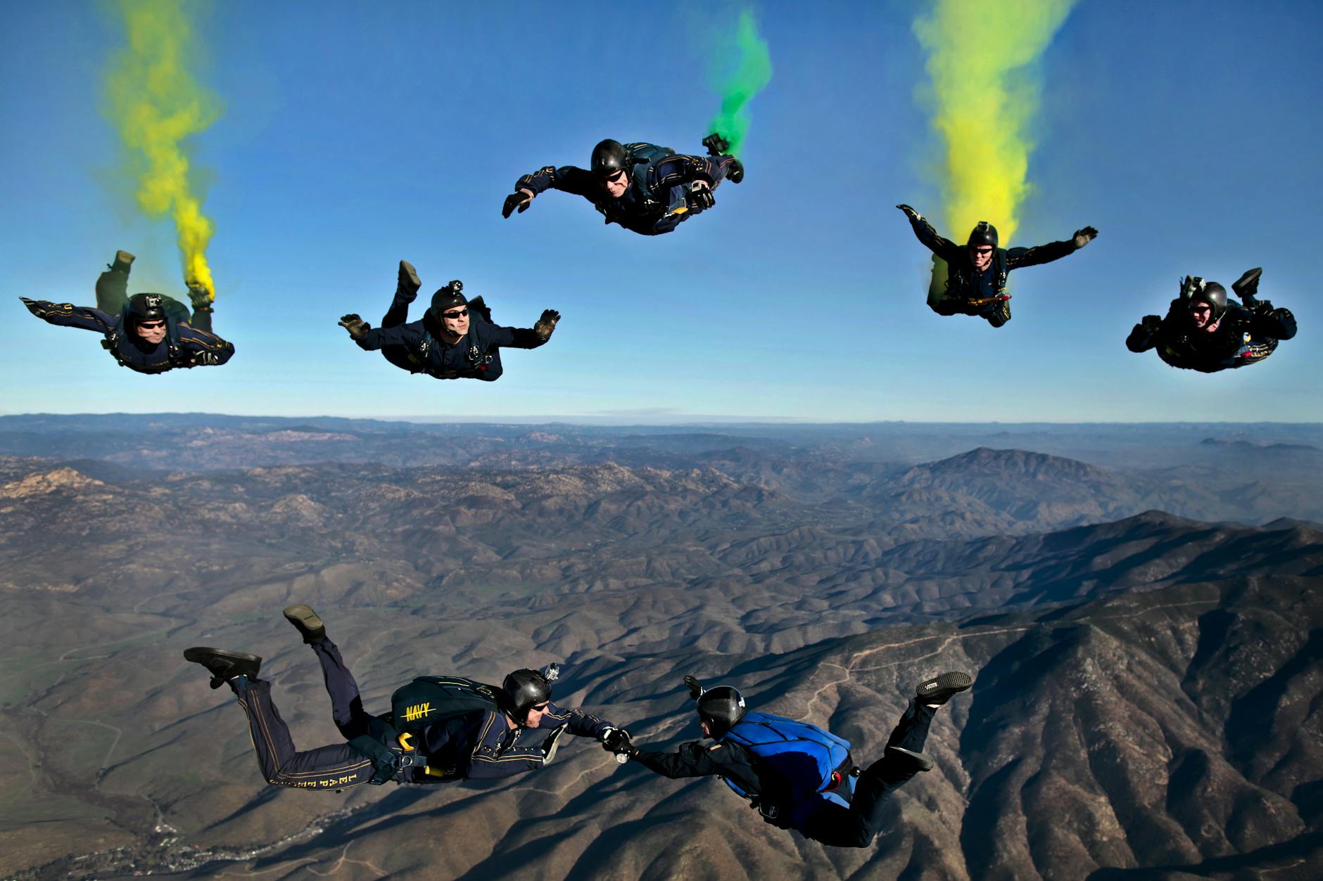Dynamic aerial shot of skydivers performing stunts with colorful smoke trails over mountainous terrain. Dynamic aerial shot of skydivers performing stunts with colorful smoke trails over mountainous terrain.