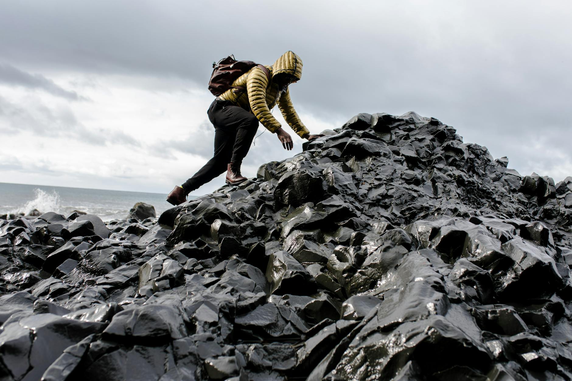 Person in yellow jacket climbs black rocky terrain by the ocean under cloudy skies. Person in yellow jacket climbs black rocky terrain by the ocean under cloudy skies.