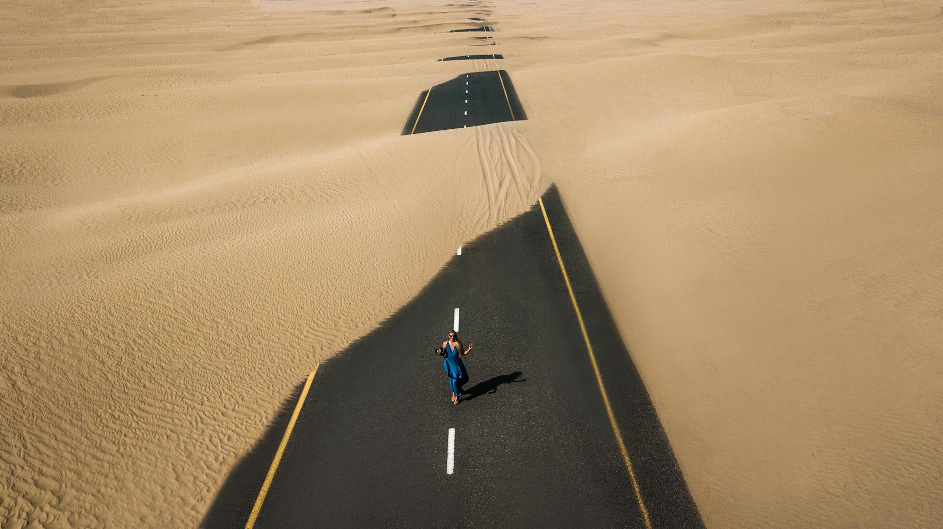 A lone woman walking on a sand-covered road in Dubai’s desert, captured from above. A lone woman walking on a sand-covered road in Dubai’s desert, captured from above.
