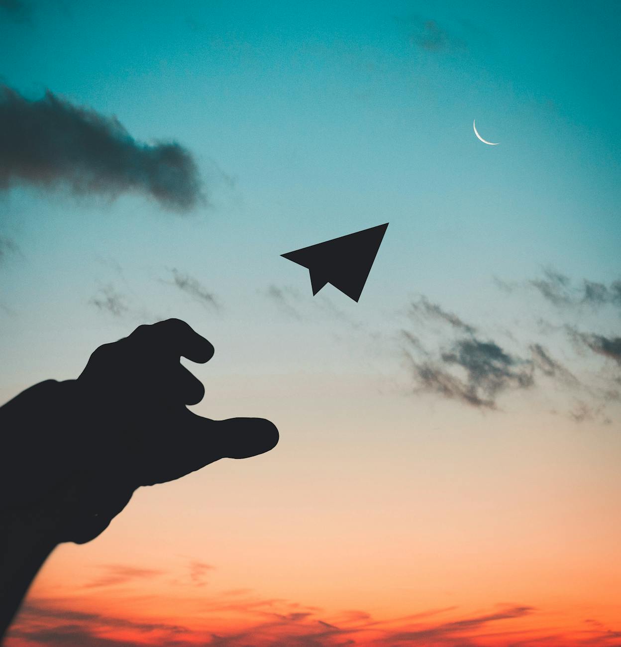 A silhouette of a hand launching a paper plane against a colorful dusk sky with a crescent moon. A silhouette of a hand launching a paper plane against a colorful dusk sky with a crescent moon.