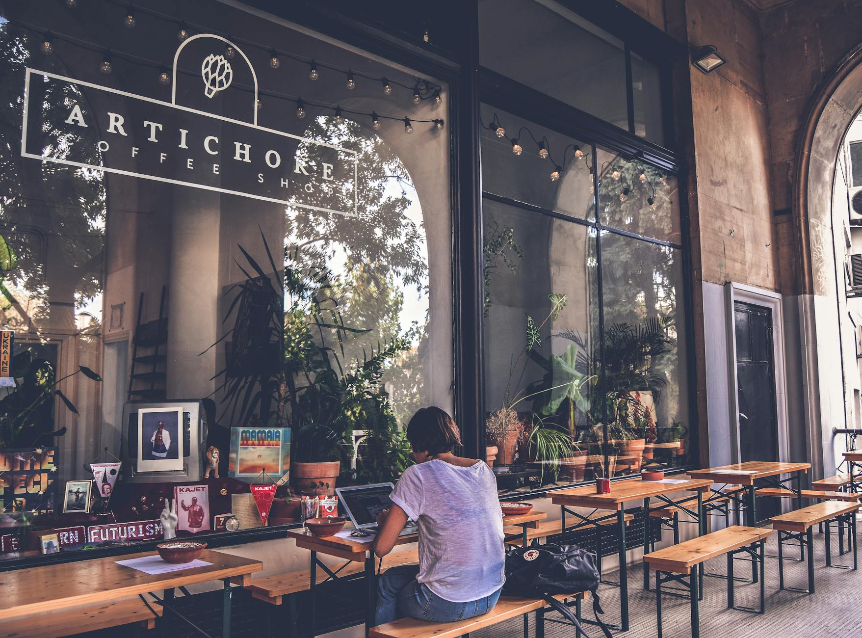 Atmospheric exterior view of a cozy cafe with wooden tables and a person working on a laptop. Atmospheric exterior view of a cozy cafe with wooden tables and a person working on a laptop.