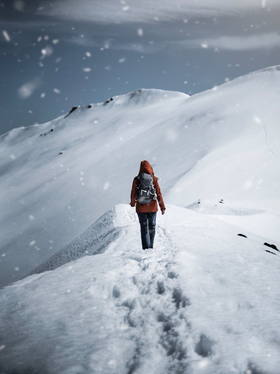 A lone hiker in a red jacket walks up a snow-covered mountain path during a snowfall. A lone hiker in a red jacket walks up a snow-covered mountain path during a snowfall.