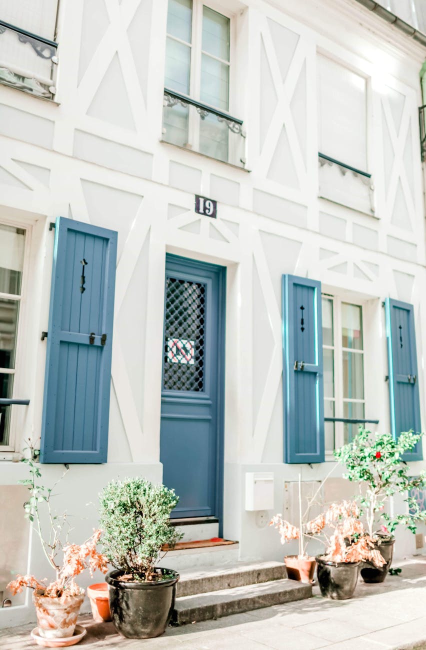 Elegant white house facade featuring blue shutters and vibrant potted plants, evoking a serene home exterior.