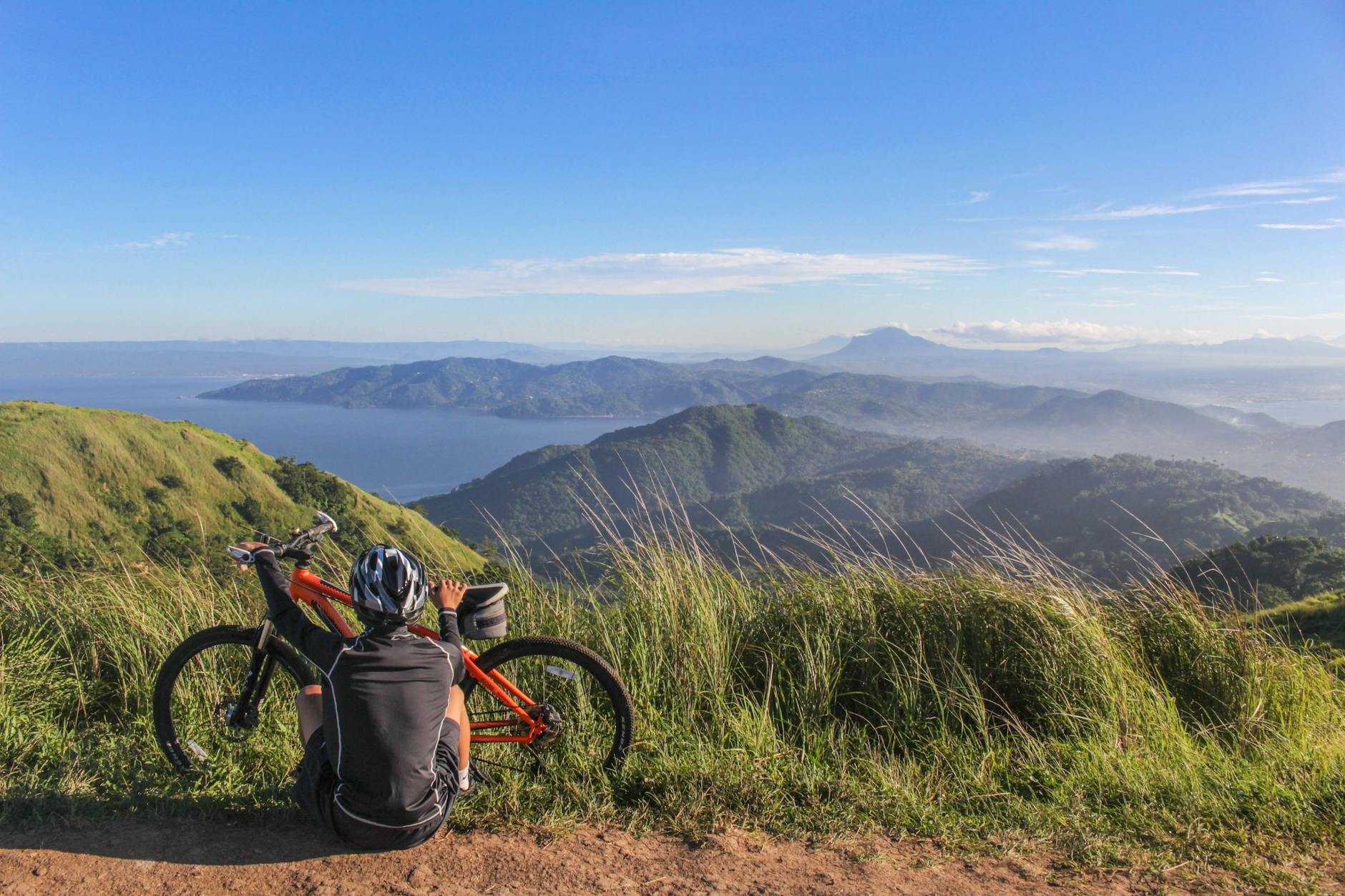 A mountain biker resting by their bike, enjoying a scenic view of lush hills and distant water under a bright blue sky. A mountain biker resting by their bike, enjoying a scenic view of lush hills and distant water under a bright blue sky.