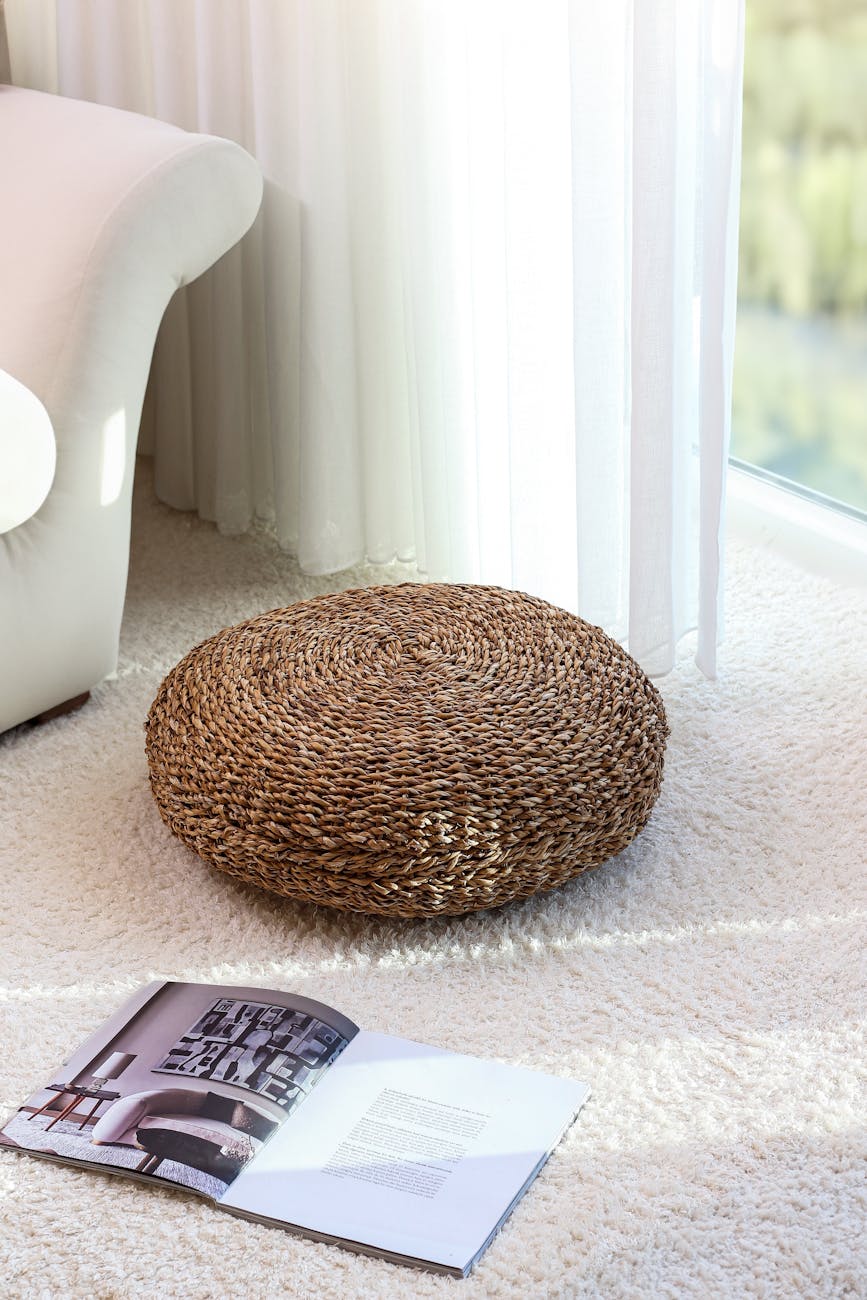 A stylish wicker pouf on shag carpet near a sofa and open magazine in bright living room. A stylish wicker pouf on shag carpet near a sofa and open magazine in bright living room.