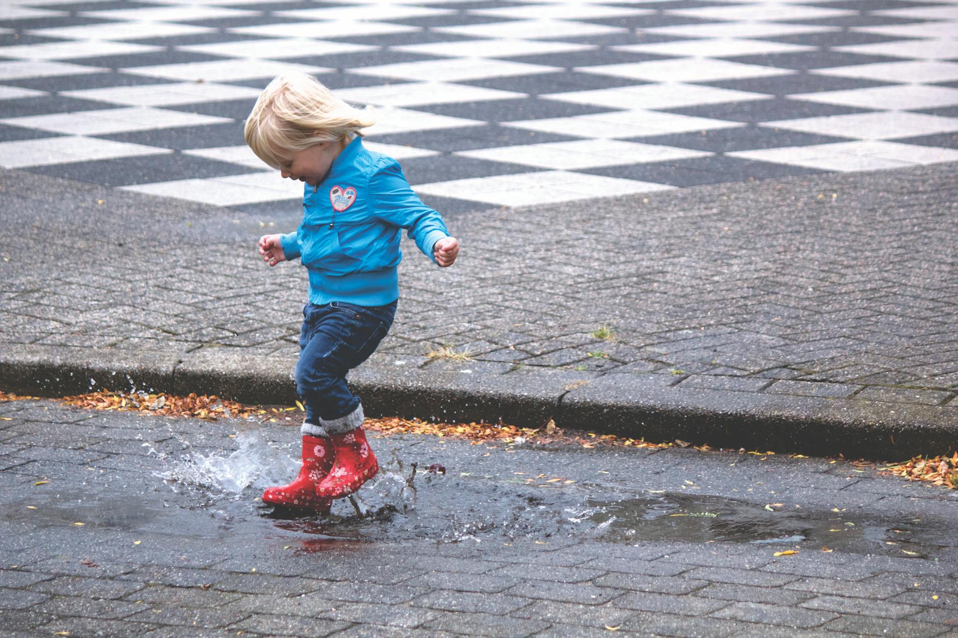 A young child joyfully splashes in a puddle wearing red boots on a rainy day.