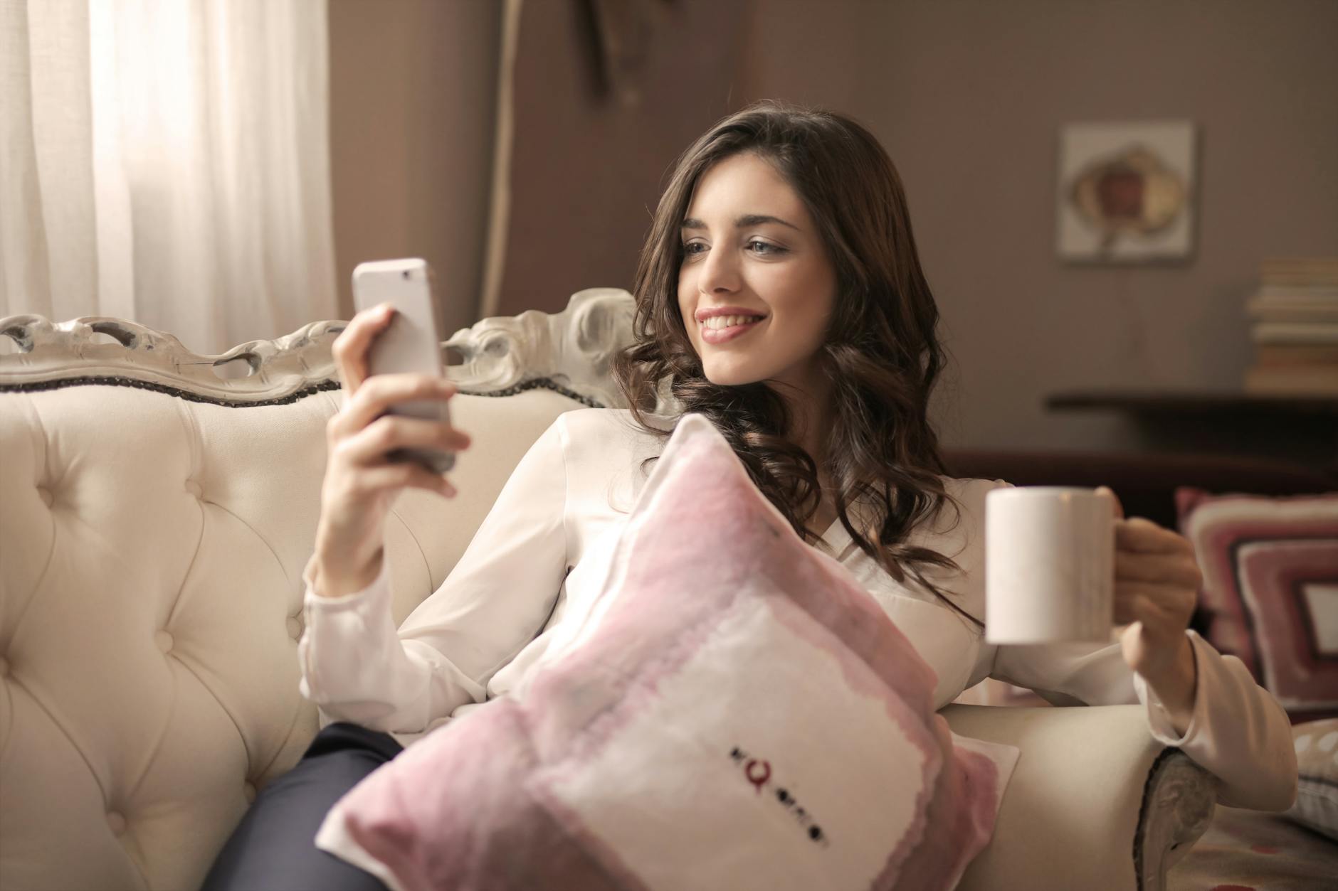 A young woman relaxing on a sofa, enjoying coffee and using her smartphone indoors. A young woman relaxing on a sofa, enjoying coffee and using her smartphone indoors.