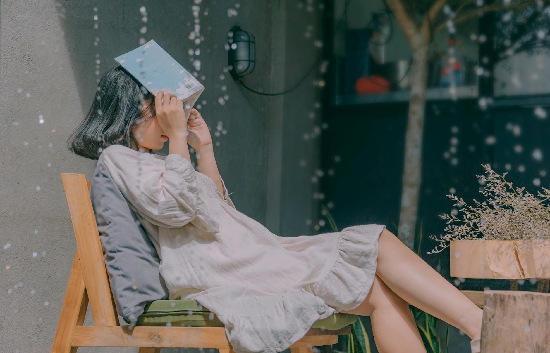 A young woman covers her face with a book while sitting outdoors on a rainy day, enjoying a peaceful moment. A young woman covers her face with a book while sitting outdoors on a rainy day, enjoying a peaceful moment.