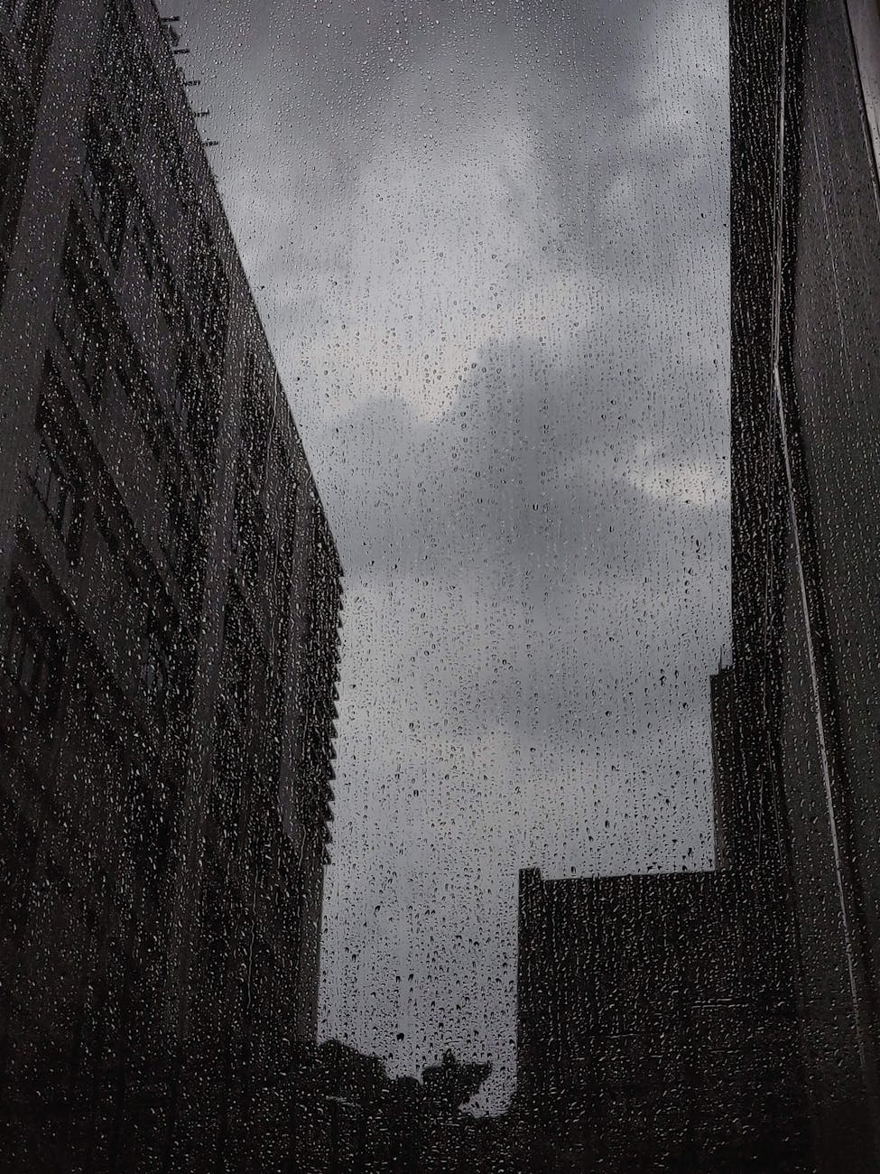 A dark urban scene with rain droplets on a window overlooking tall buildings. A dark urban scene with rain droplets on a window overlooking tall buildings.