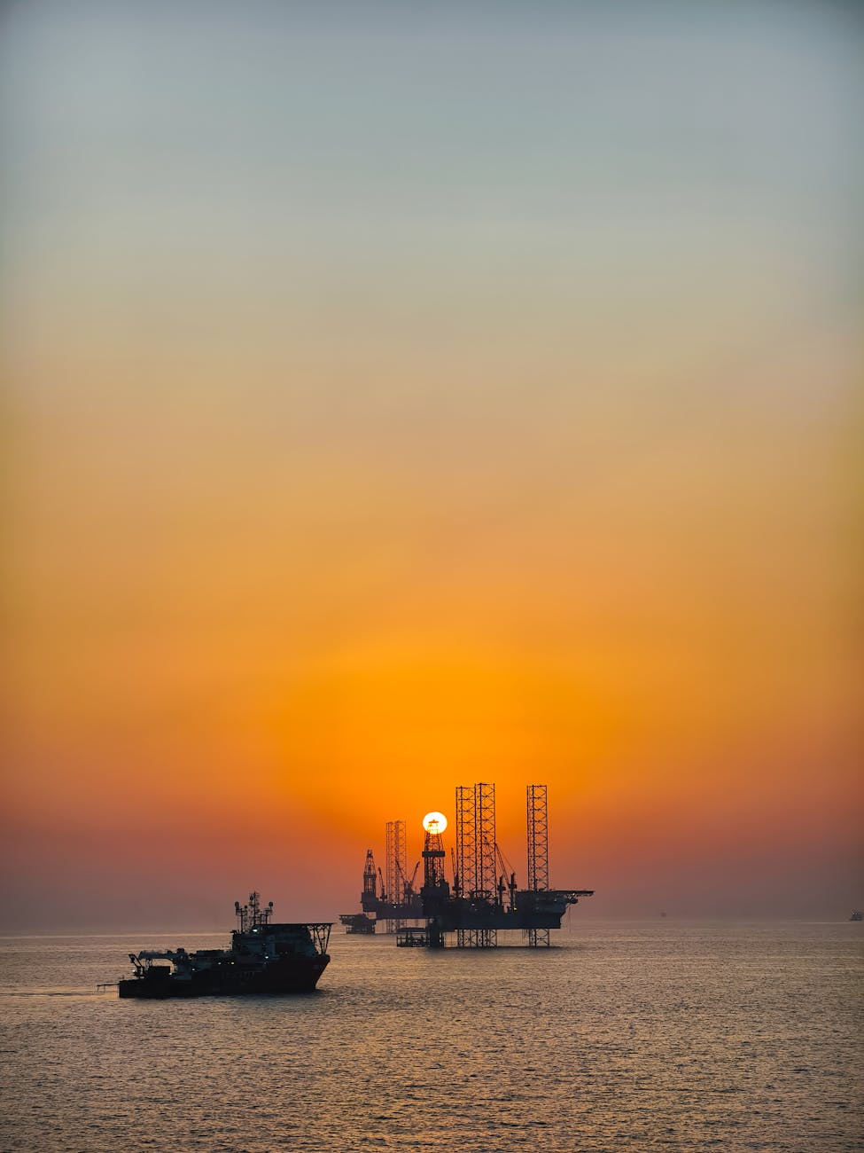 Offshore oil rigs silhouetted against a stunning orange sunset over calm seas.
