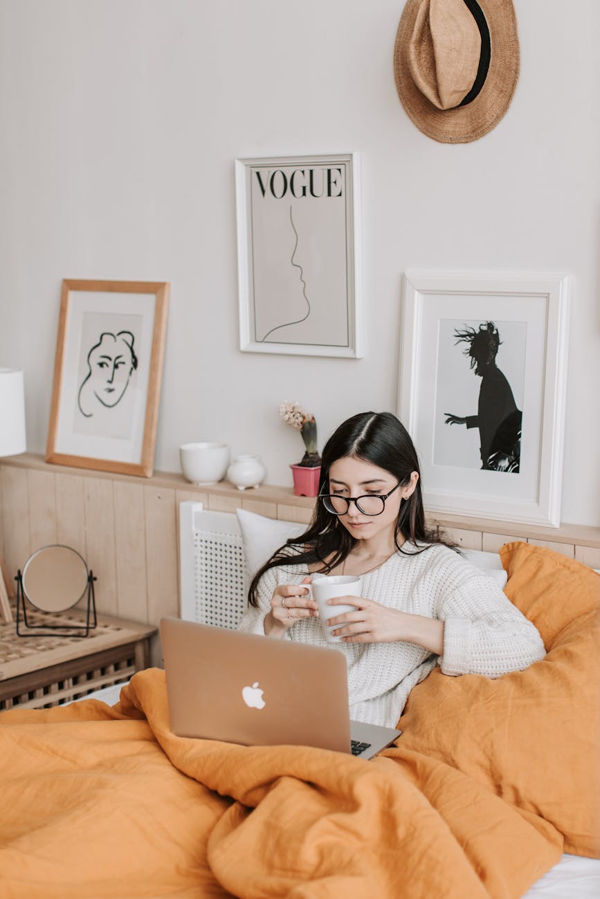 A woman relaxing in bed with coffee and laptop, surrounded by art and comfort. A woman relaxing in bed with coffee and laptop, surrounded by art and comfort.