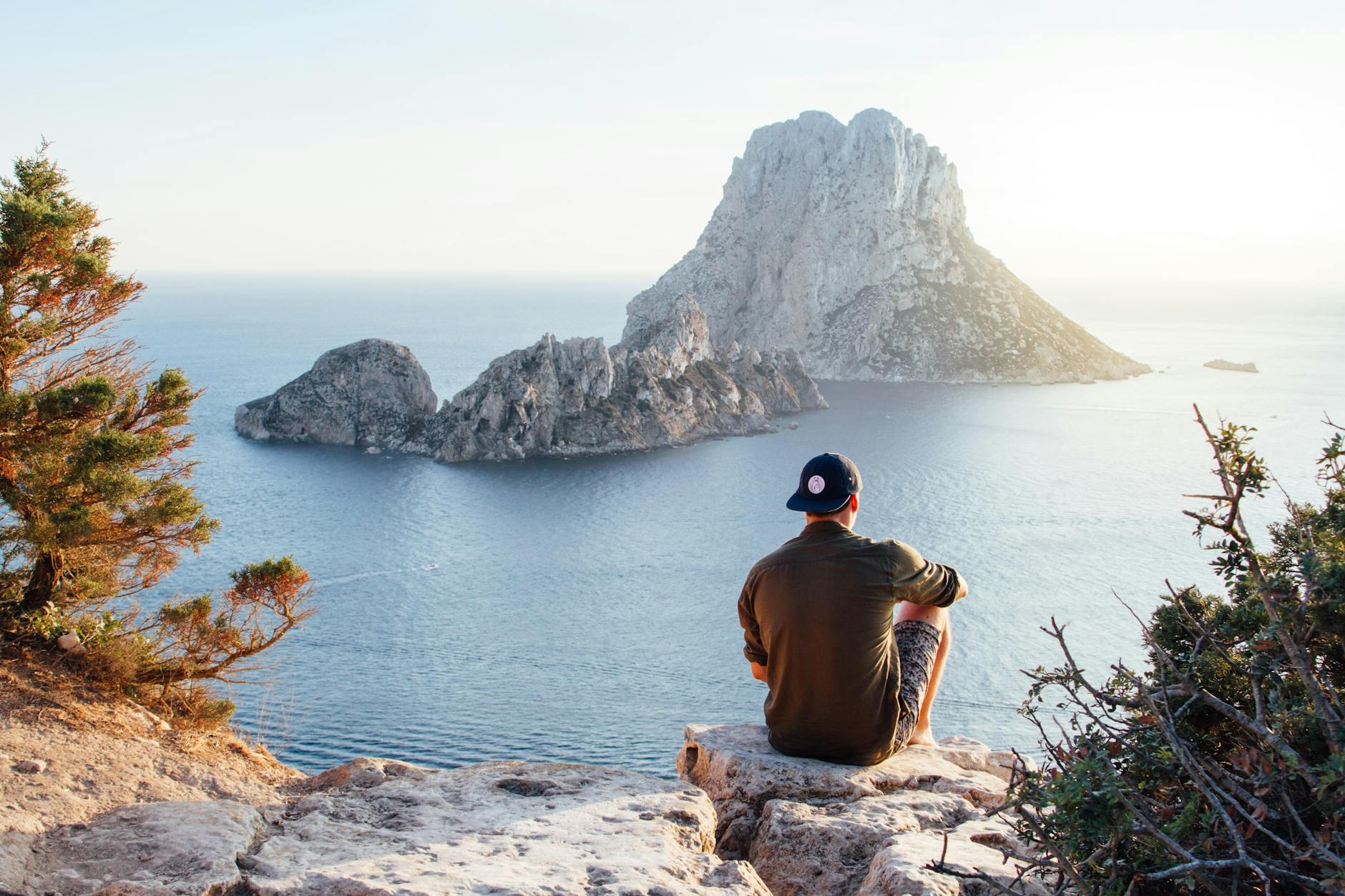 Man enjoys a scenic view of Es Vedrà at sunset from a cliff in San Juan Bautista, providing a perfect summer escape. Man enjoys a scenic view of Es Vedrà at sunset from a cliff in San Juan Bautista, providing a perfect summer escape.