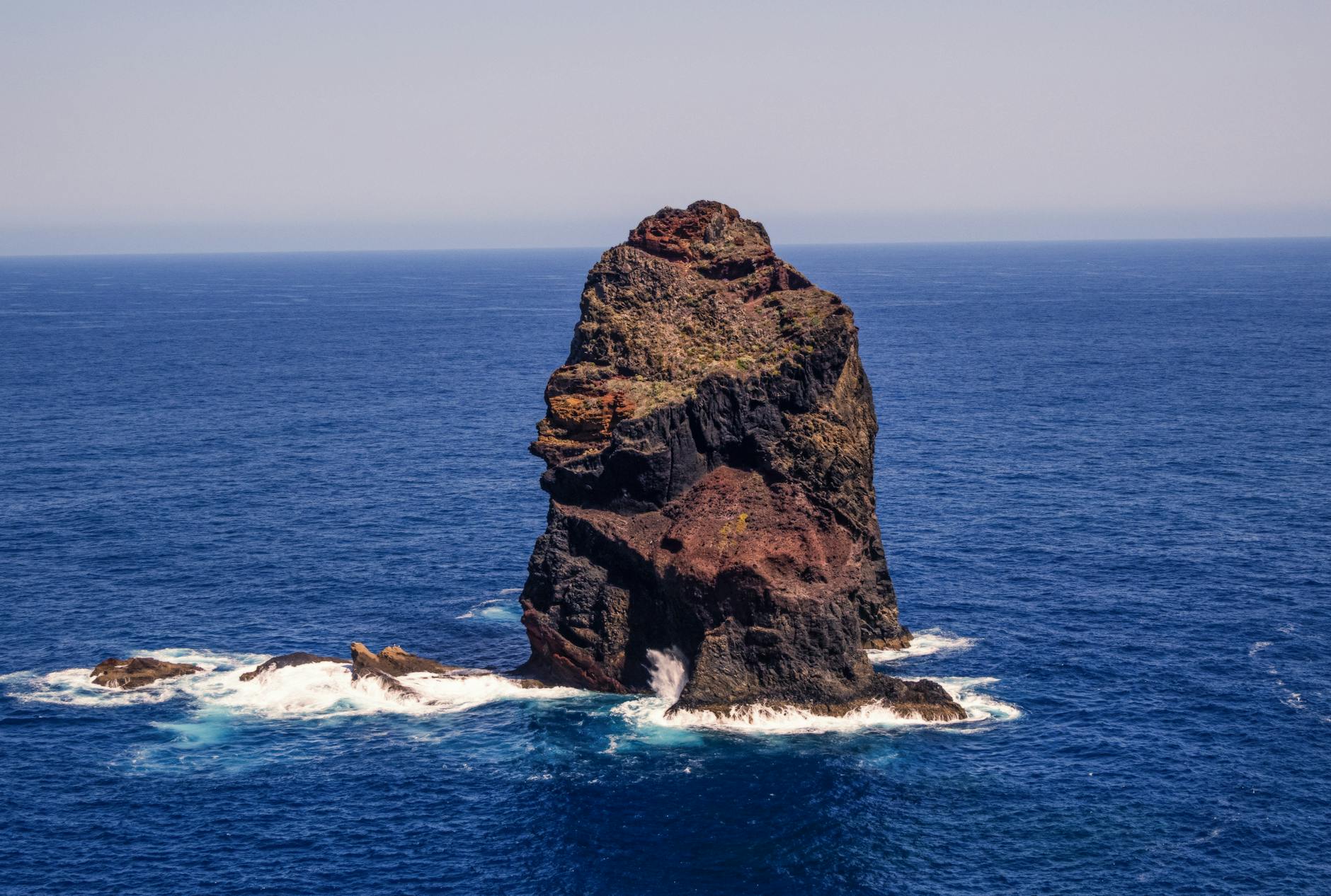 A striking oceanic rock formation stands alone amidst vibrant blue sea waves under a clear sky.