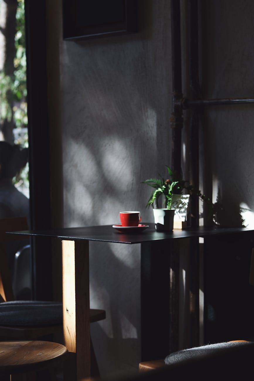 A quiet cafe table lit by dappled sunlight, featuring a single red coffee cup. A quiet cafe table lit by dappled sunlight, featuring a single red coffee cup.
