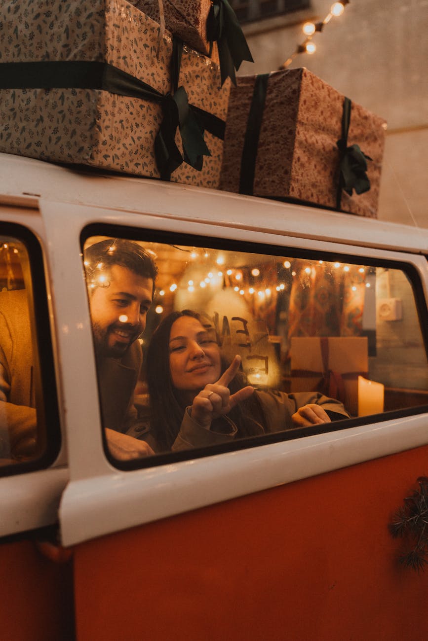 A joyful couple in a decorated van surrounded by festive holiday lights and stacked gifts, embodying holiday spirit. A joyful couple in a decorated van surrounded by festive holiday lights and stacked gifts, embodying holiday spirit.
