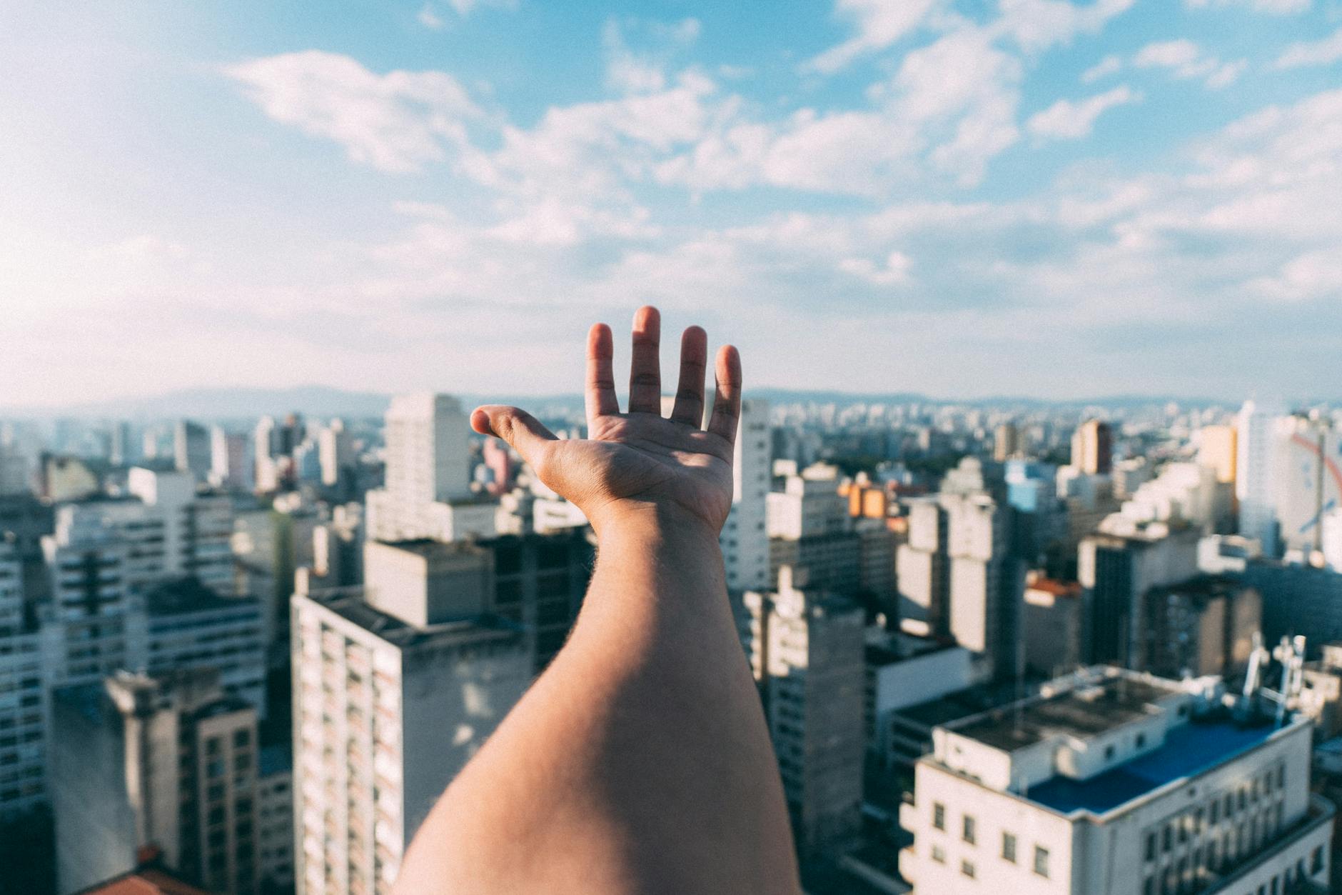 An outstretched hand reaching towards a sunny cityscape, capturing the vibrant skyline and skyscrapers. An outstretched hand reaching towards a sunny cityscape, capturing the vibrant skyline and skyscrapers.