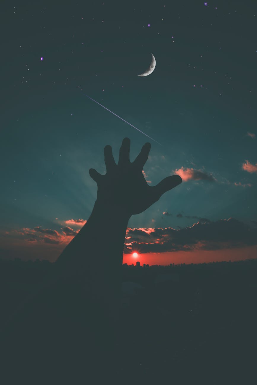 Silhouette of a hand reaching towards the crescent moon and stars in a dramatic dusk sky.