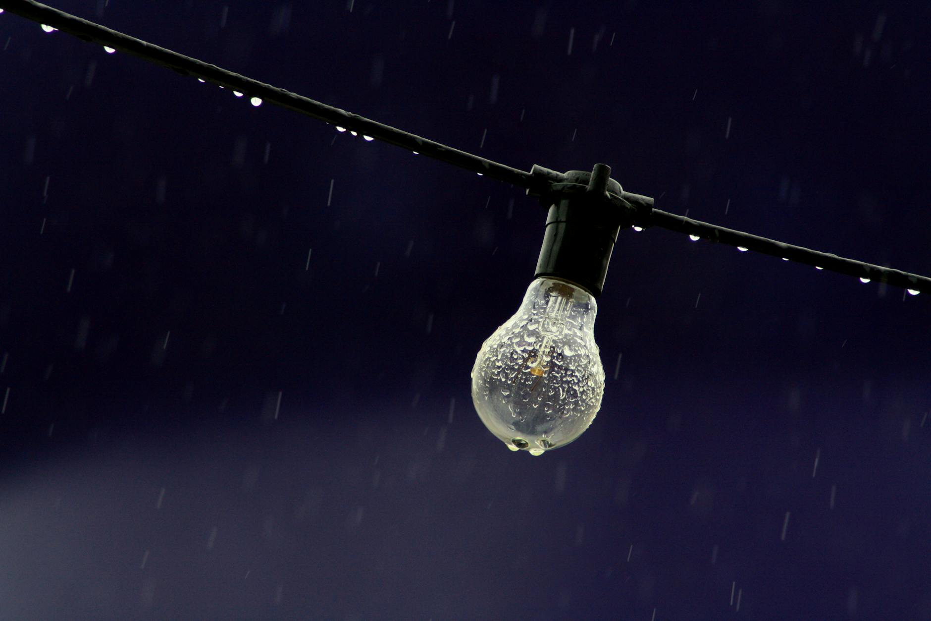 A clear lightbulb with raindrops hanging on a wire against a deep blue sky.