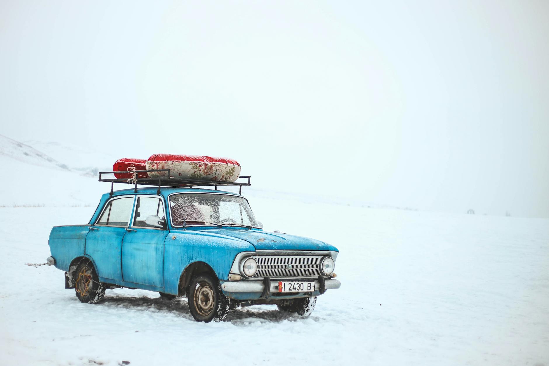 A vintage blue car with luggage on top amidst a snowy landscape in Kyrgyzstan.