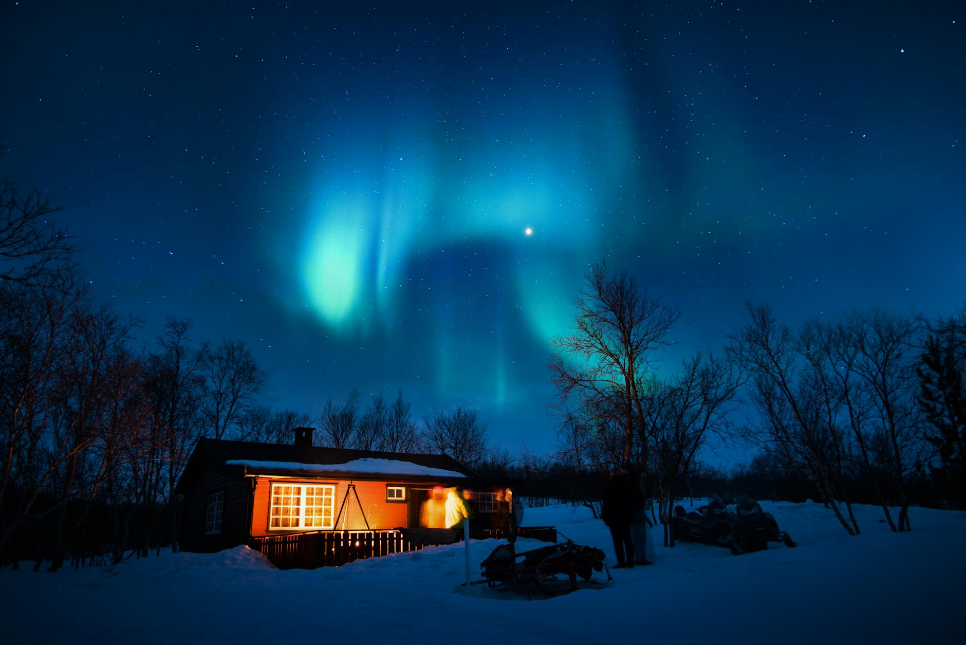 Cabin illuminated under a stunning aurora borealis and starry winter night sky.