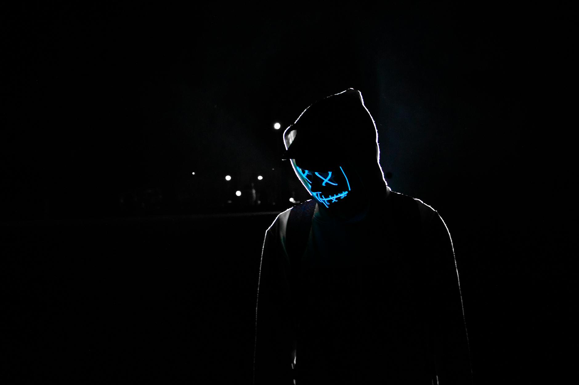 Silhouette of a person in a hoodie wearing a glowing LED mask against a dark background. Silhouette of a person in a hoodie wearing a glowing LED mask against a dark background.