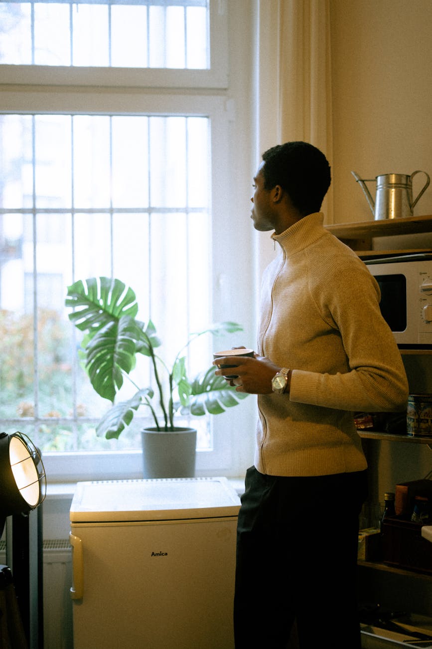 Calm man holding a cup and gazing out the window indoors, surrounded by cozy decor.