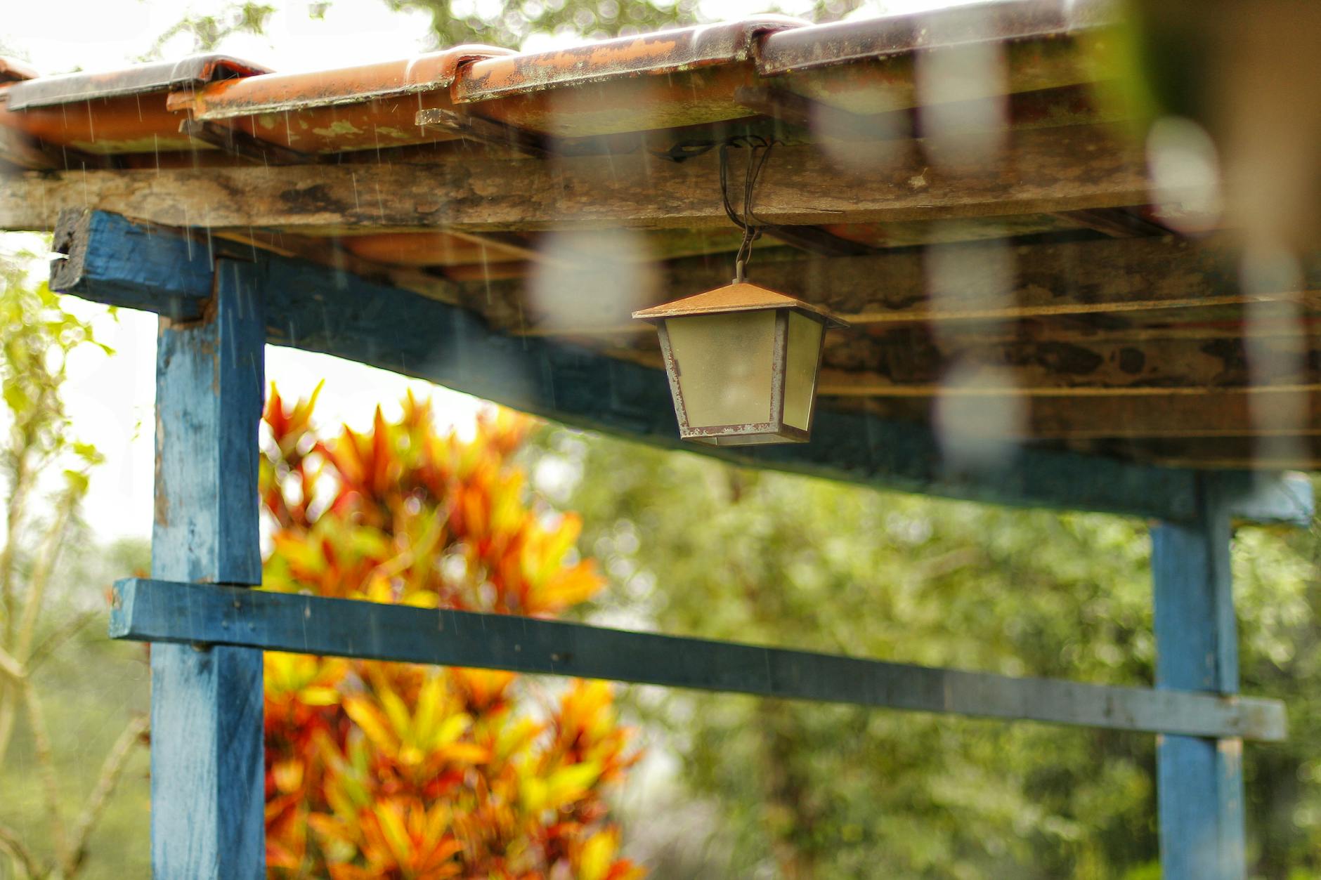 Rustic wooden structure with a hanging lamp under a rainy sky, providing a cozy outdoor setting.